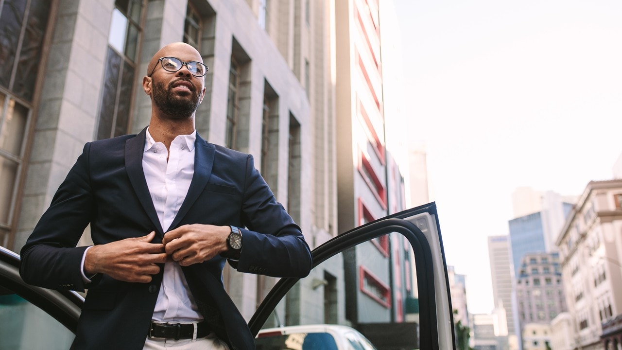 Portrait of a handsome young businessman getting down from car on road and buttoning his jacket. African man standing by his car in city.