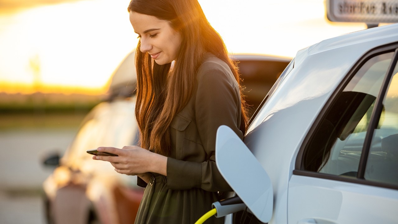 Side view waist up shot of a young woman in a dress using her mobile phone while waiting for her white electric car to charge during sunset