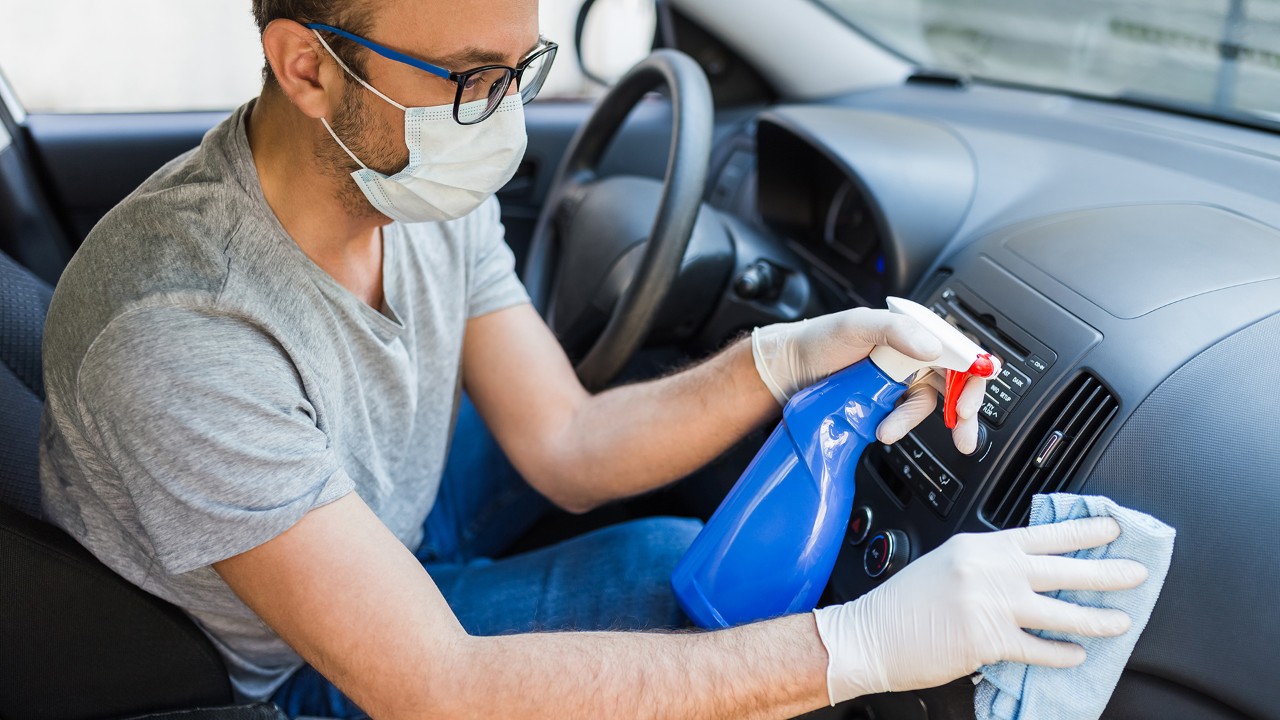 Driver with face surgical mask using disinfectant and microfiber cloth to clean car interior.