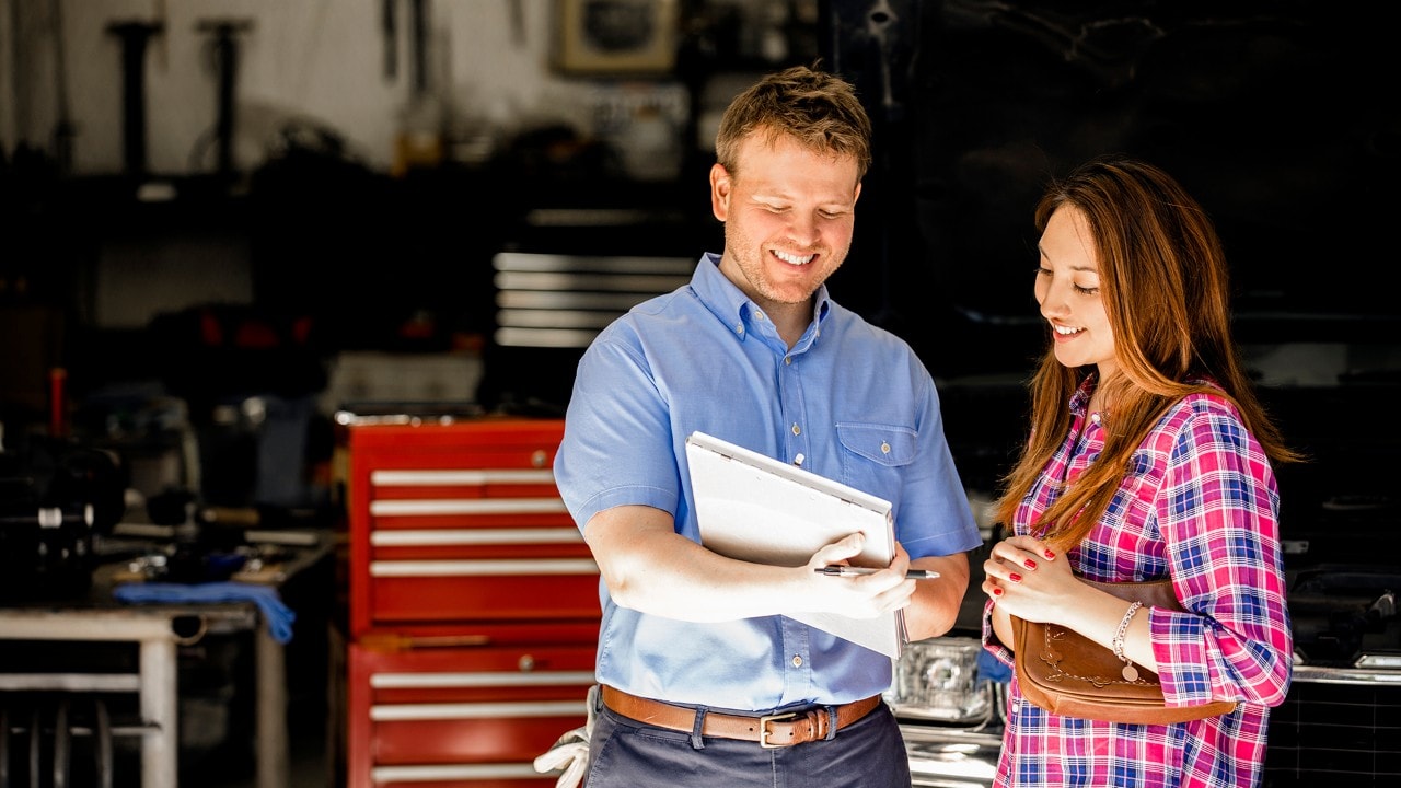 Happy customer!  A smiling Latin descent woman discusses automobile repair invoice with an auto mechanic in a repair shop.  She is discussing the vehicle's repairs with the mechanic, who is explaining her service.  Toolbox, workshop background. Unidentifiable, SUV-style vehicle.